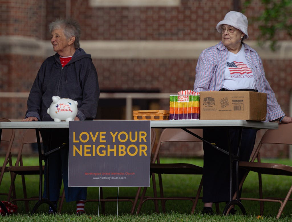 Two elderly women volunteer at a community event with a 'Love Your Neighbor' sign.